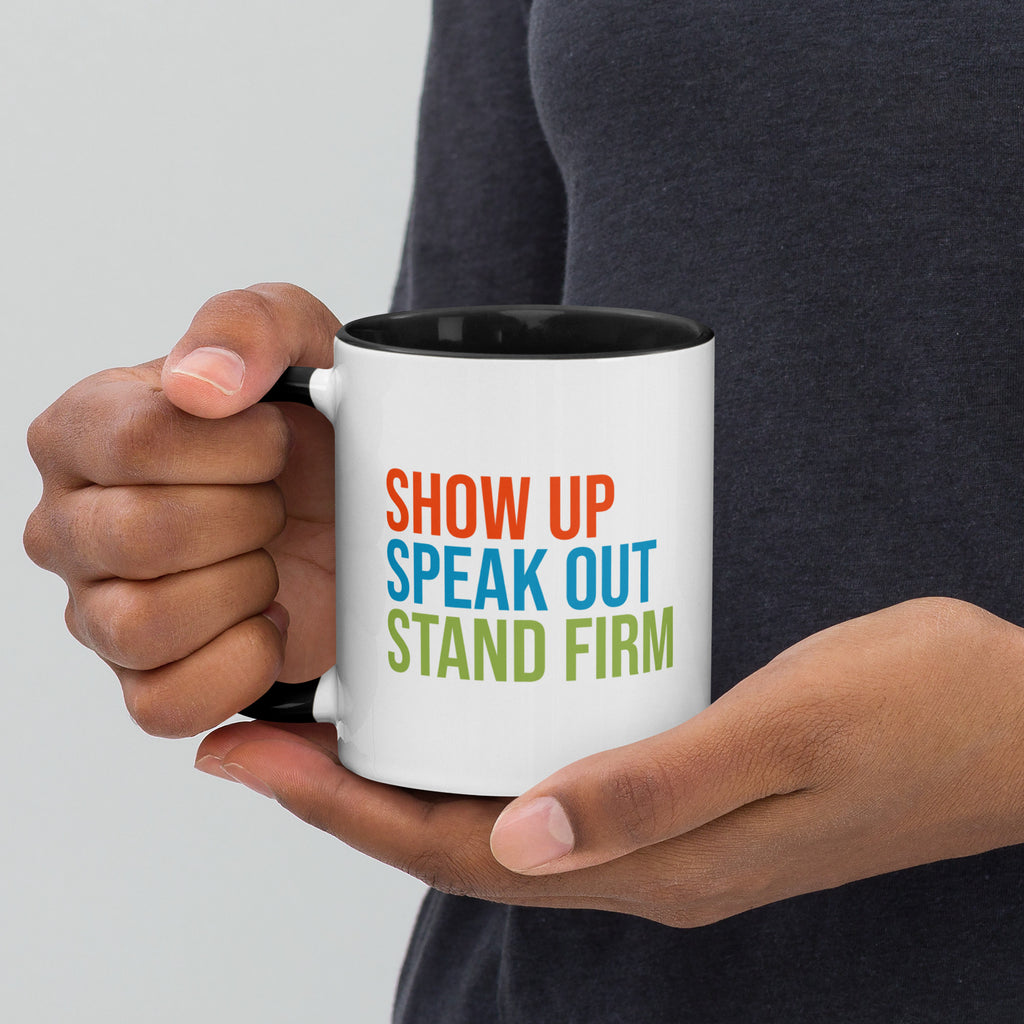 Coffee mug with black handle and inside with text on one side that reads "show up, speak out, stand firm." The other side, not shown in photo, reads "democracy doesn't defend itself."