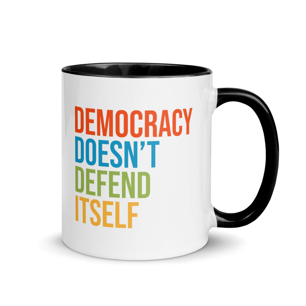 Coffee mug with black handle and inside with text on one side that reads "democracy doesn't defend itself." The other side, not shown in photo, reads "show up, speak out, stand firm."