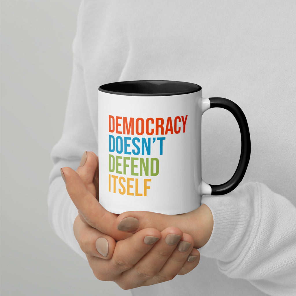 Coffee mug with black handle and inside with text on one side that reads "democracy doesn't defend itself." The other side, not shown in photo, reads "show up, speak out, stand firm."