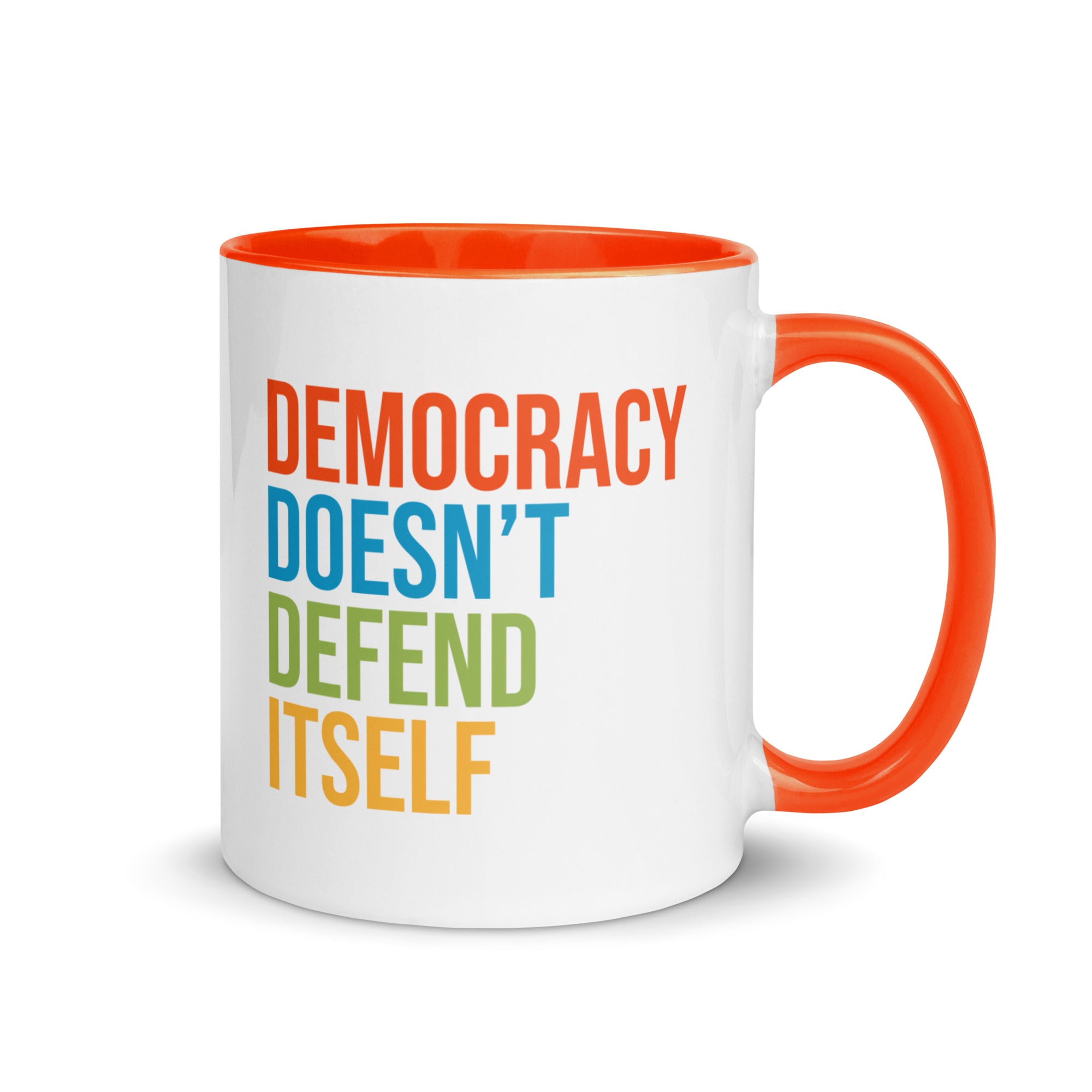 Coffee mug with orange handle and inside with text on one side that reads "democracy doesn't defend itself." The other side, not shown in photo, reads "show up, speak out, stand firm."