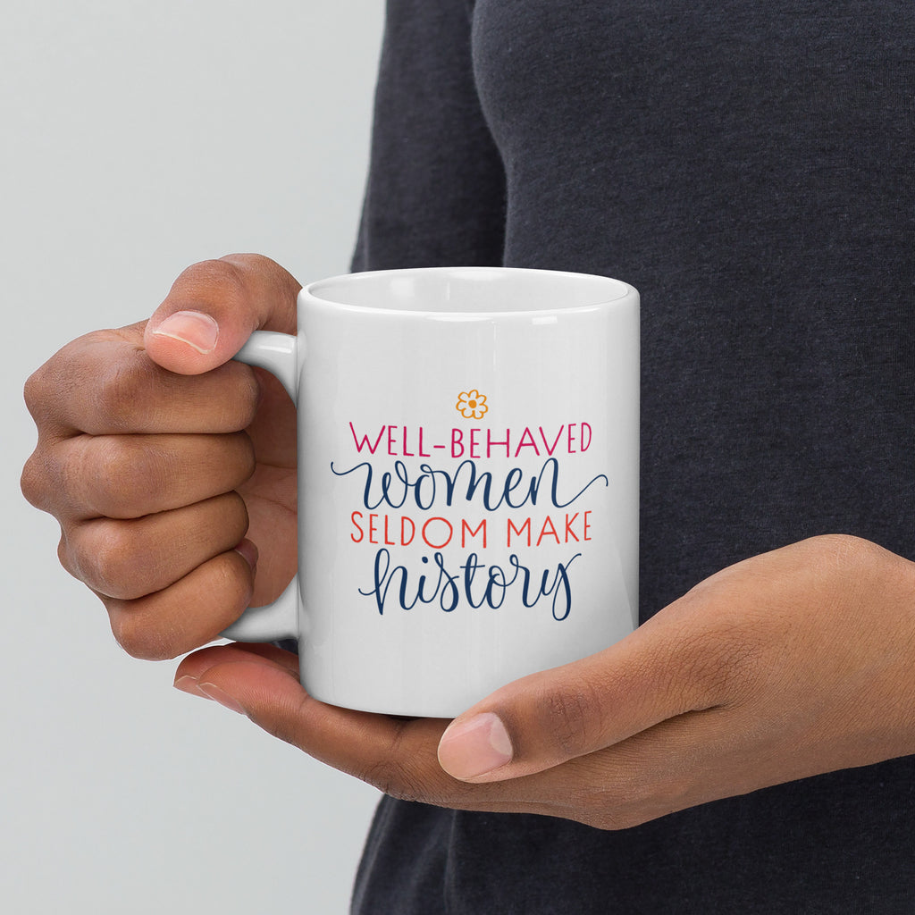 Person holding a white mug with colorful text that reads "well-behaved women seldom make history" against a neutral background