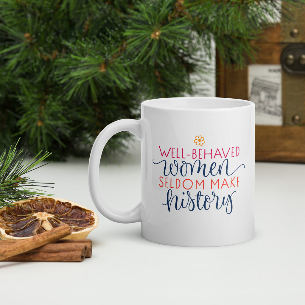 White mug with a motivational quote "well-behaved women seldom make history" on a surface with decorative forest elements.