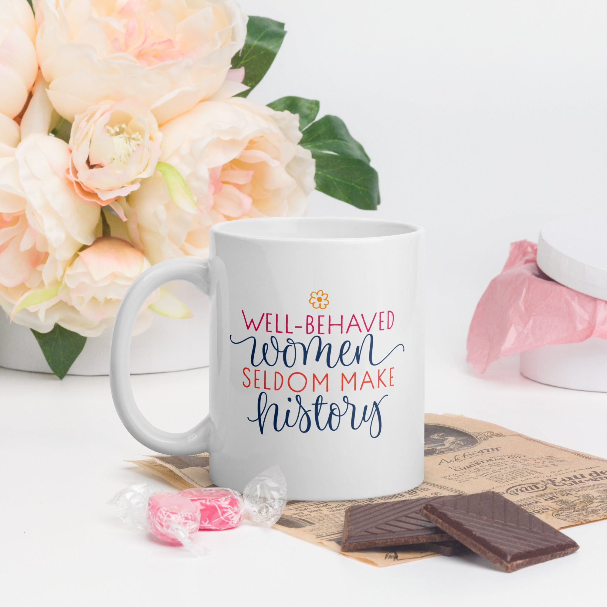 White mug with 'Well-behaved women seldom make history' text, flowers, and chocolate on a white background