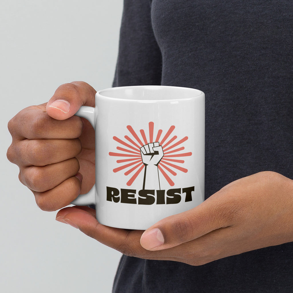 Person holding coffee mug with handle on left. Mugh has a raised fist graphic in front of a coral burst, and bold retro-styled text that says "resist."