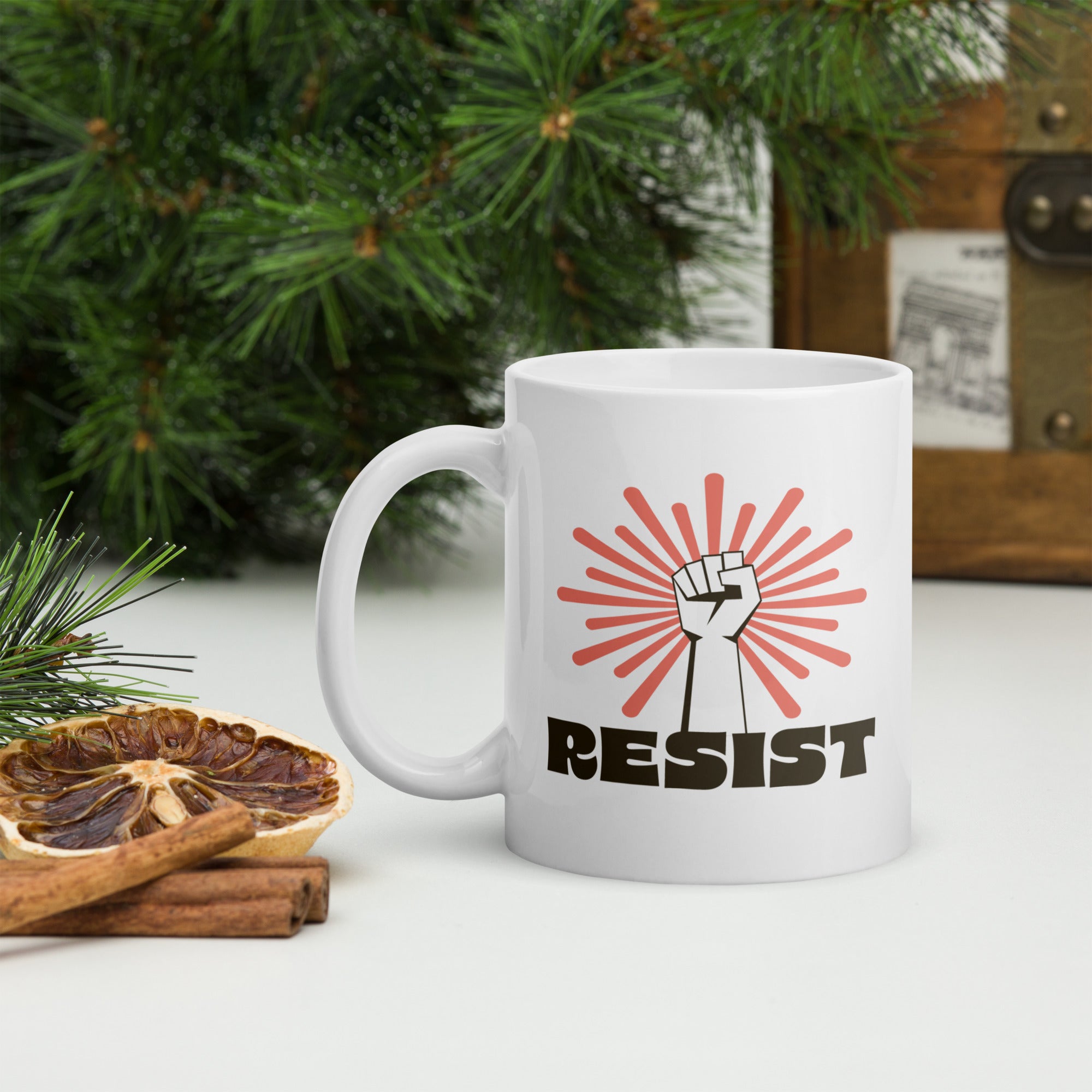 Coffee mug in a rustic tree scene. Mug has a raised fist graphic in front of a coral burst, and bold retro-styled text that says "resist."