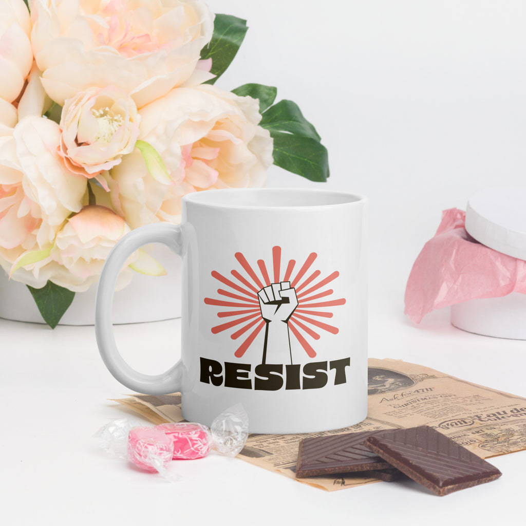 Coffee mug in a scene with Valentine's day flowers and candy. Mug has a raised fist graphic in front of a coral burst, and bold retro-styled text that says "resist."