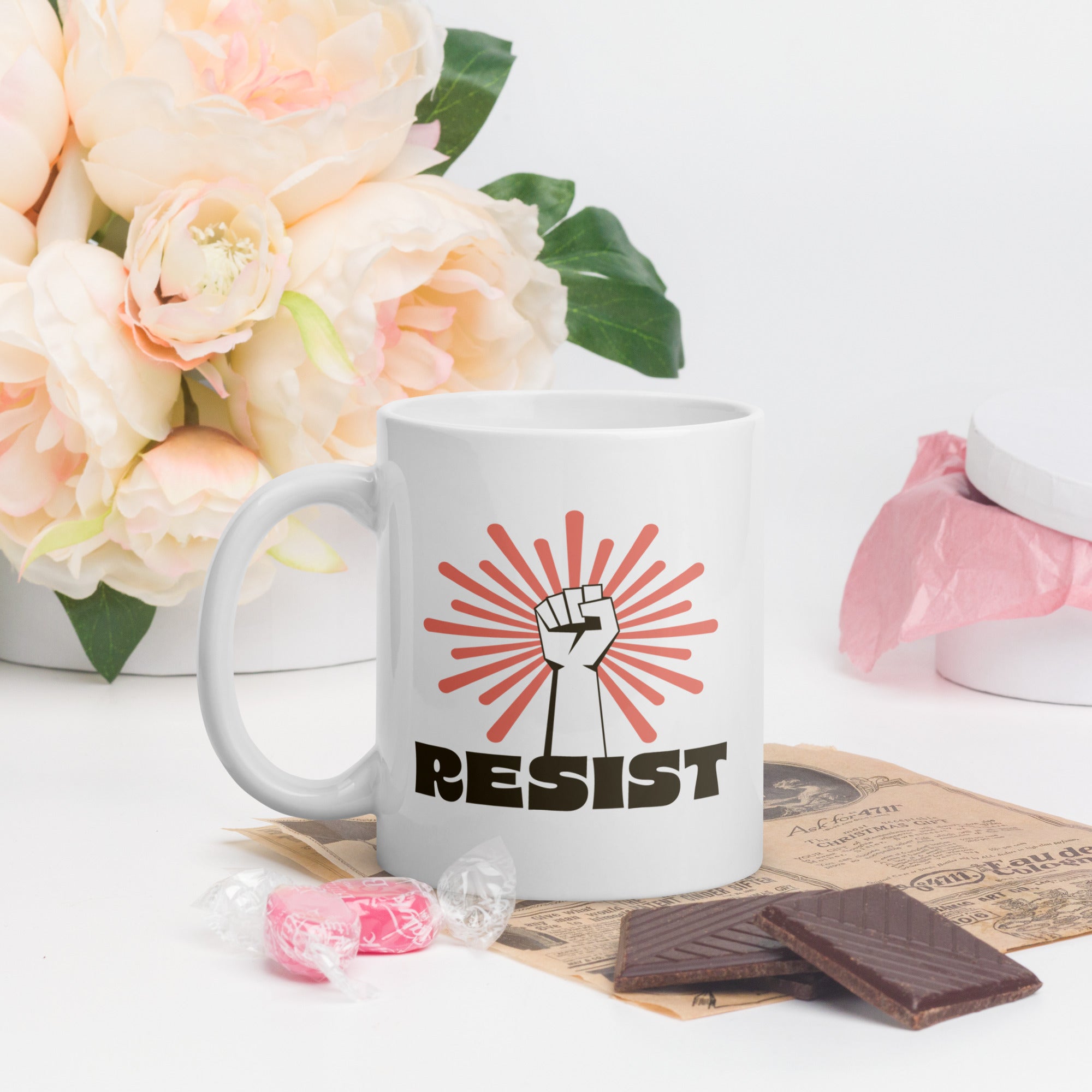 Coffee mug in a scene with Valentine's day flowers and candy. Mug has a raised fist graphic in front of a coral burst, and bold retro-styled text that says "resist."