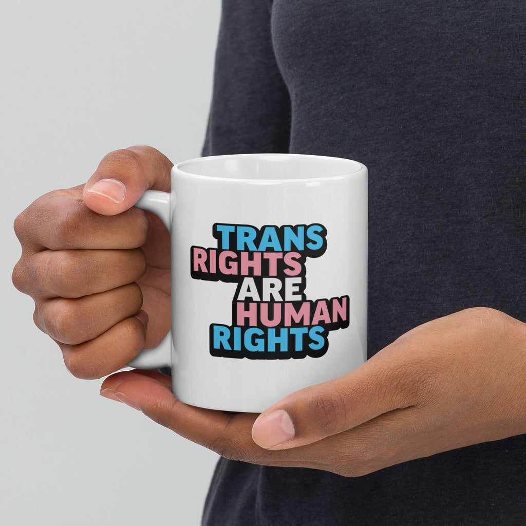 Person holding white ceramic mug with bold text “Trans Rights Are Human Rights” in blue, pink, and black colors representing the transgender pride flag, with handle on the left.