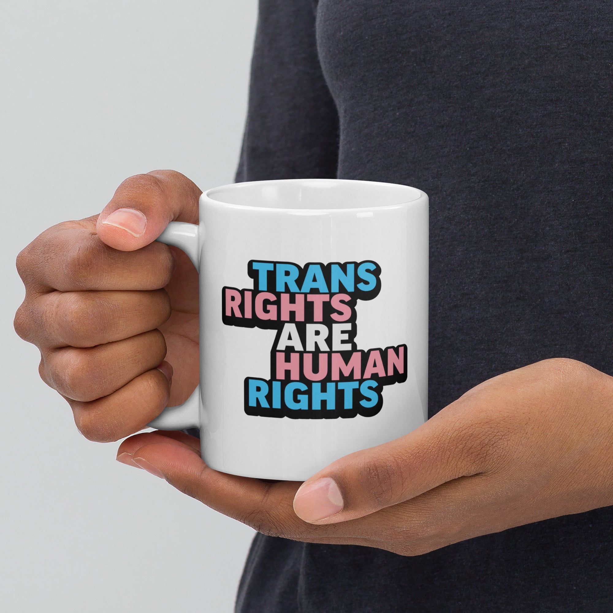 Person holding white ceramic mug with bold text “Trans Rights Are Human Rights” in blue, pink, and black colors representing the transgender pride flag, with handle on the left.