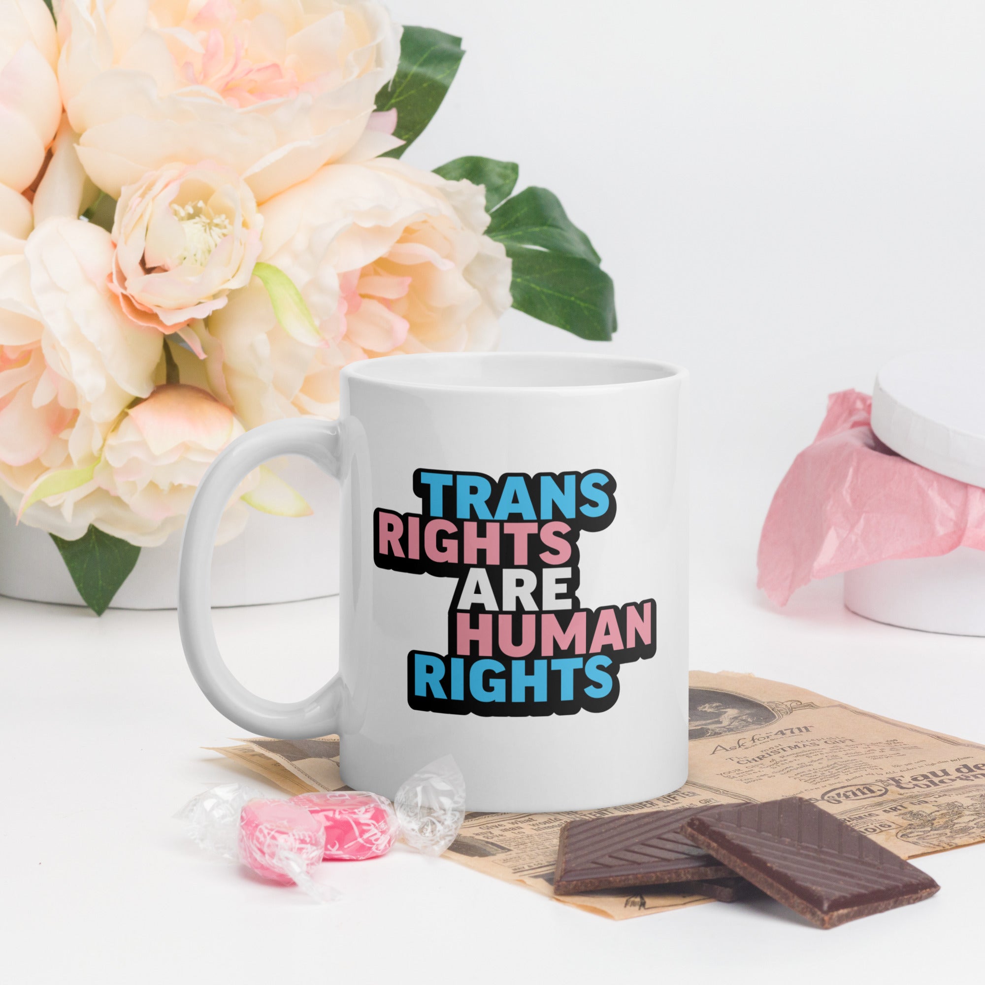 White ceramic mug with bold text “Trans Rights Are Human Rights” in blue, pink, and black colors representing the transgender pride flag, on a table surrounded by valentine's candy.