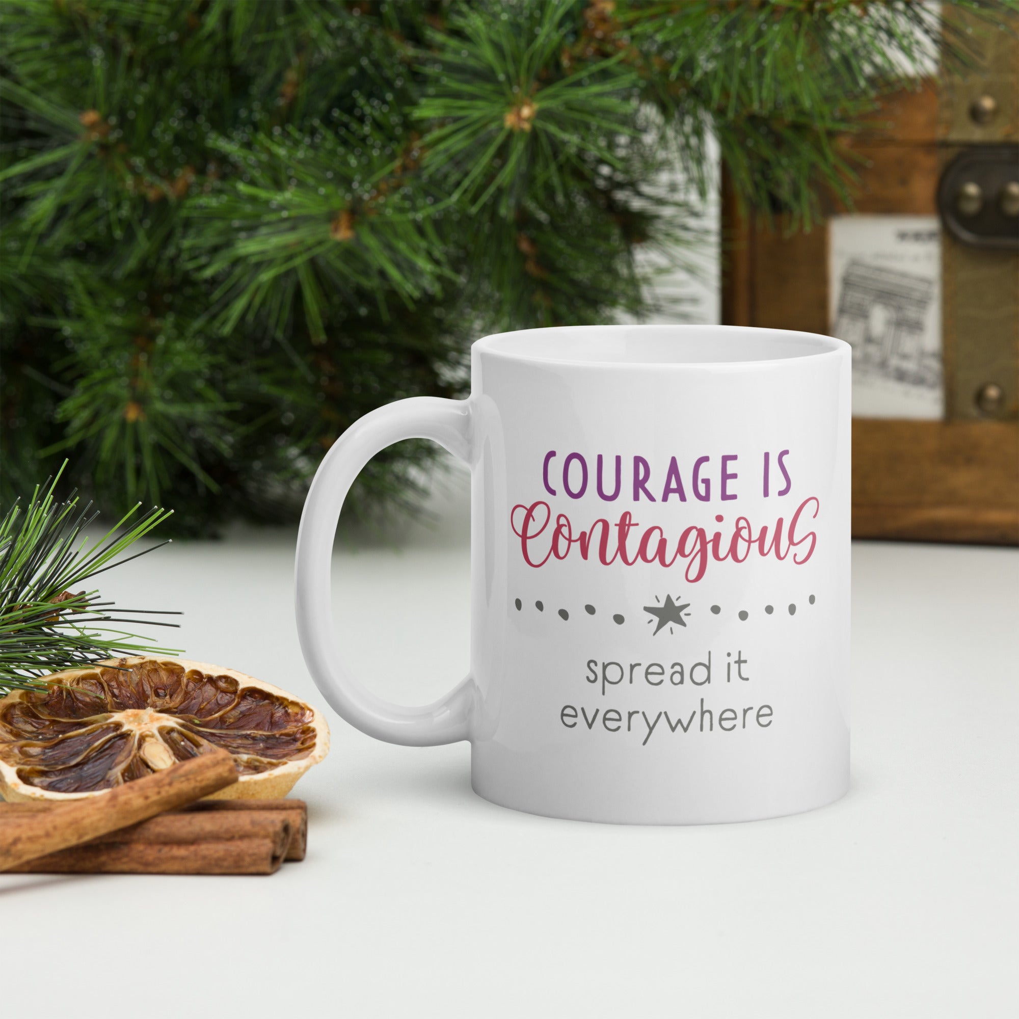 Mug with styled text graphic that reads "courage is contagious – spread it everywhere", on table with forest greenersy surrounding it.