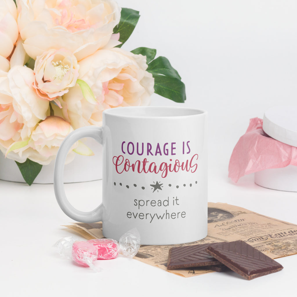 Mug with styled text graphic that reads "courage is contagious – spread it everywhere", on a table with valentine's day candy and flowers.