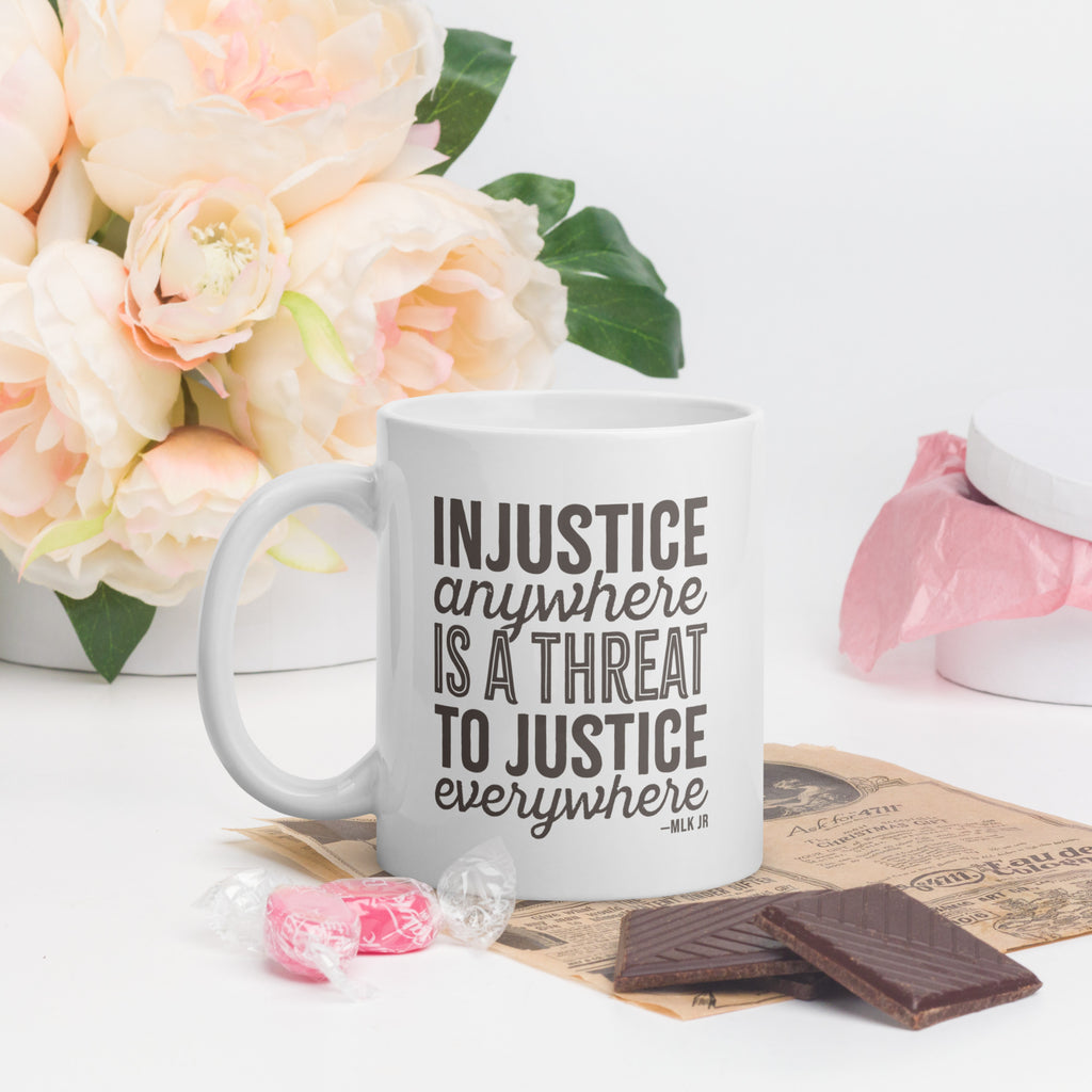 White ceramic mug with bold text graphic reading “injustice anywhere is a threat to justice everywhere,” a quote from MLK Junior from Letters from Birmingham Jail, on a table with flowers and candy.