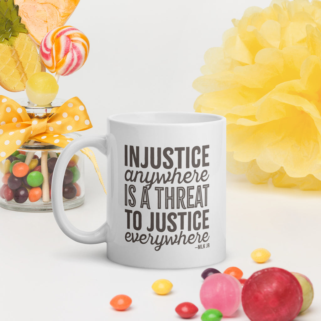 White ceramic mug with bold text graphic reading “injustice anywhere is a threat to justice everywhere,” a quote from MLK Junior from Letters from Birmingham Jail, on a table with colorful candy.