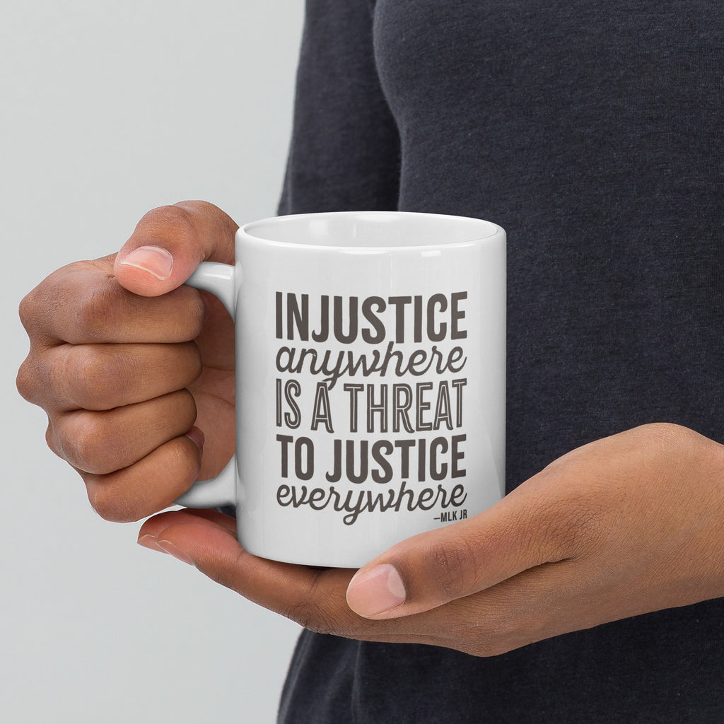 Person holding white ceramic mug showing left side with bold text graphic reading “injustice anywhere is a threat to justice everywhere,” a quote from MLK Junior from Letters from Birmingham Jail.