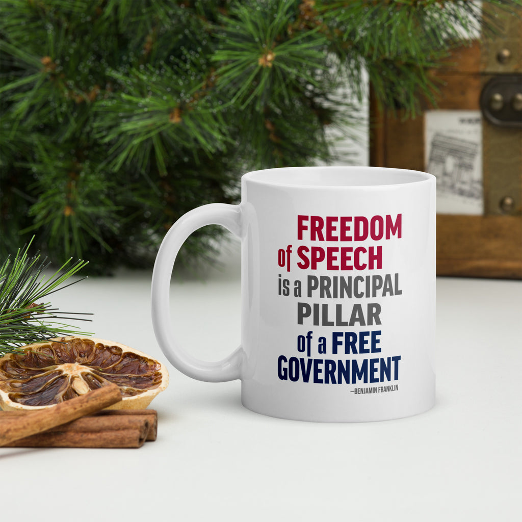 White ceramic mug with bold patriotic red, blue, and grey text that reads "Freedom of speech is a principal pillar of a free government. – Ben Franklin." Sitting on a table top with forest greenery.