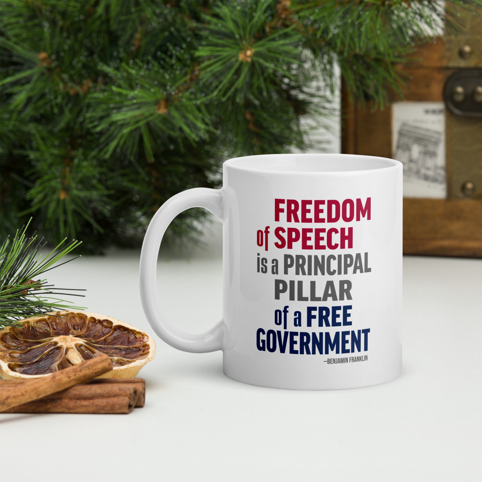 White ceramic mug with bold patriotic red, blue, and grey text that reads "Freedom of speech is a principal pillar of a free government. – Ben Franklin." Sitting on a table top with forest greenery.