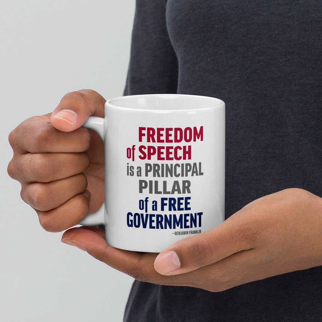 Person holding white ceramic mug with bold patriotic red, blue, and grey text that reads "Freedom of speech is a principal pillar of a free government. – Ben Franklin."
