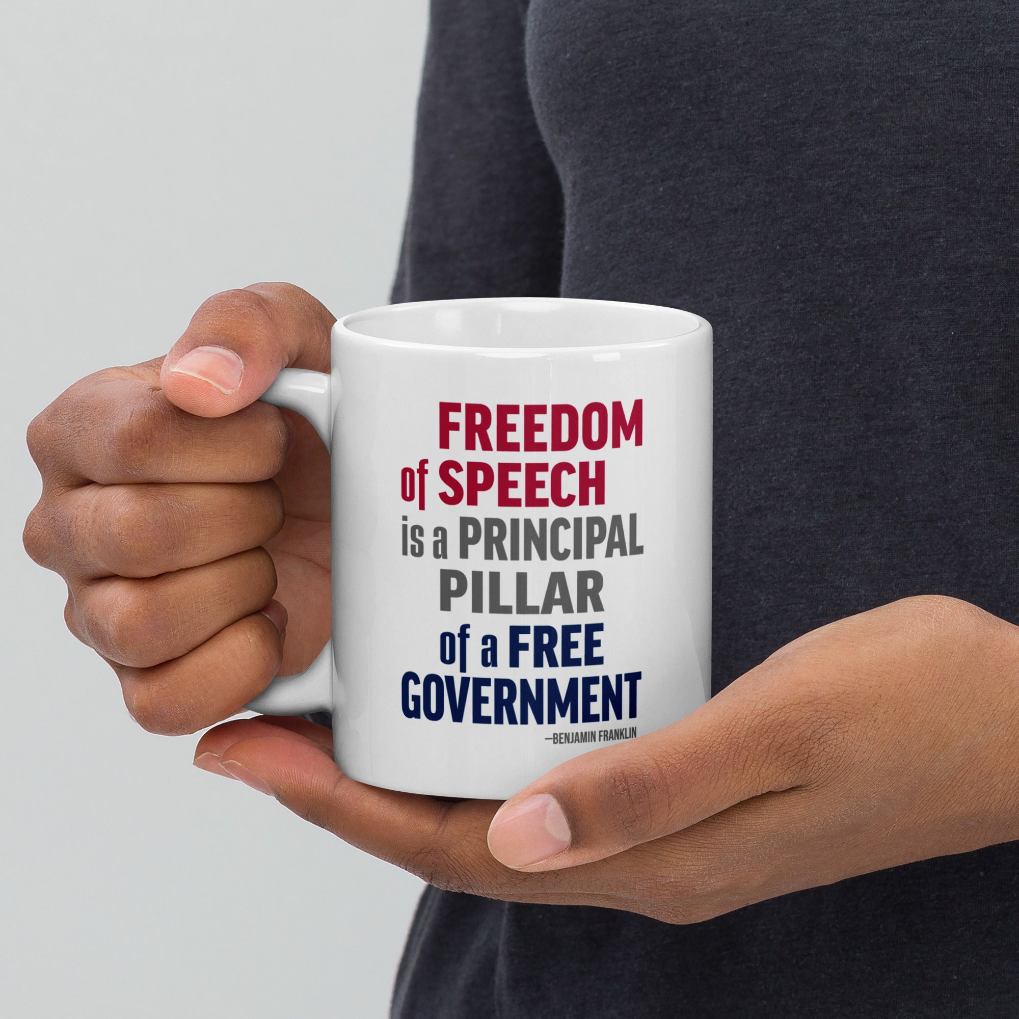 Person holding white ceramic mug with bold patriotic red, blue, and grey text that reads "Freedom of speech is a principal pillar of a free government. – Ben Franklin."