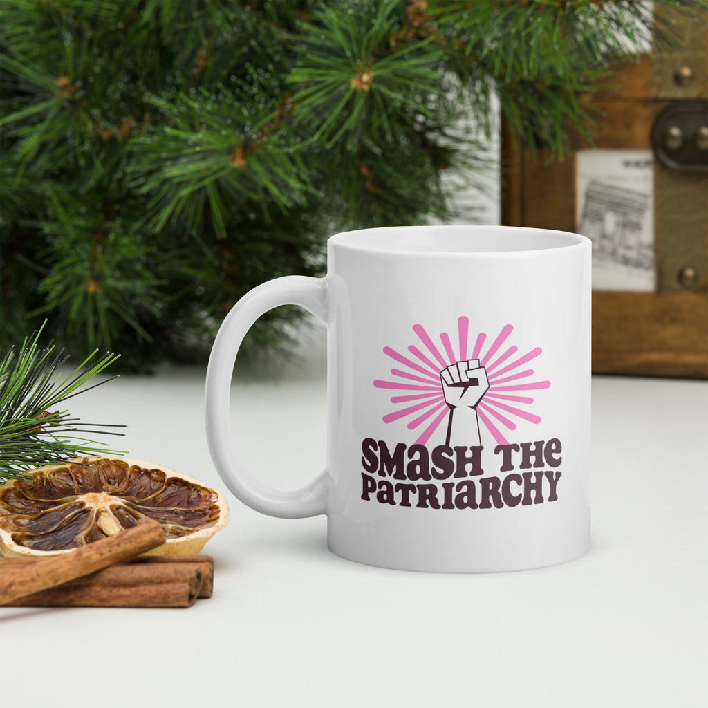 Coffee mug that says "smash the patriarchy" below a raised fist centered in front of pink rays, with handle on right, on a tabletop with forest greenery.