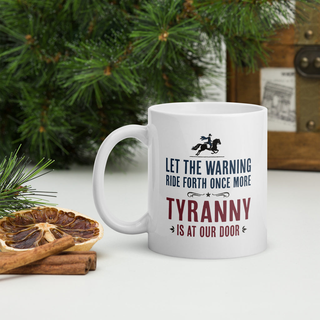 White ceramic coffee mug with a historical-style design reading “Let the warning ride forth once more — Tyranny is at our door,” on a tabletop with forest greenery.