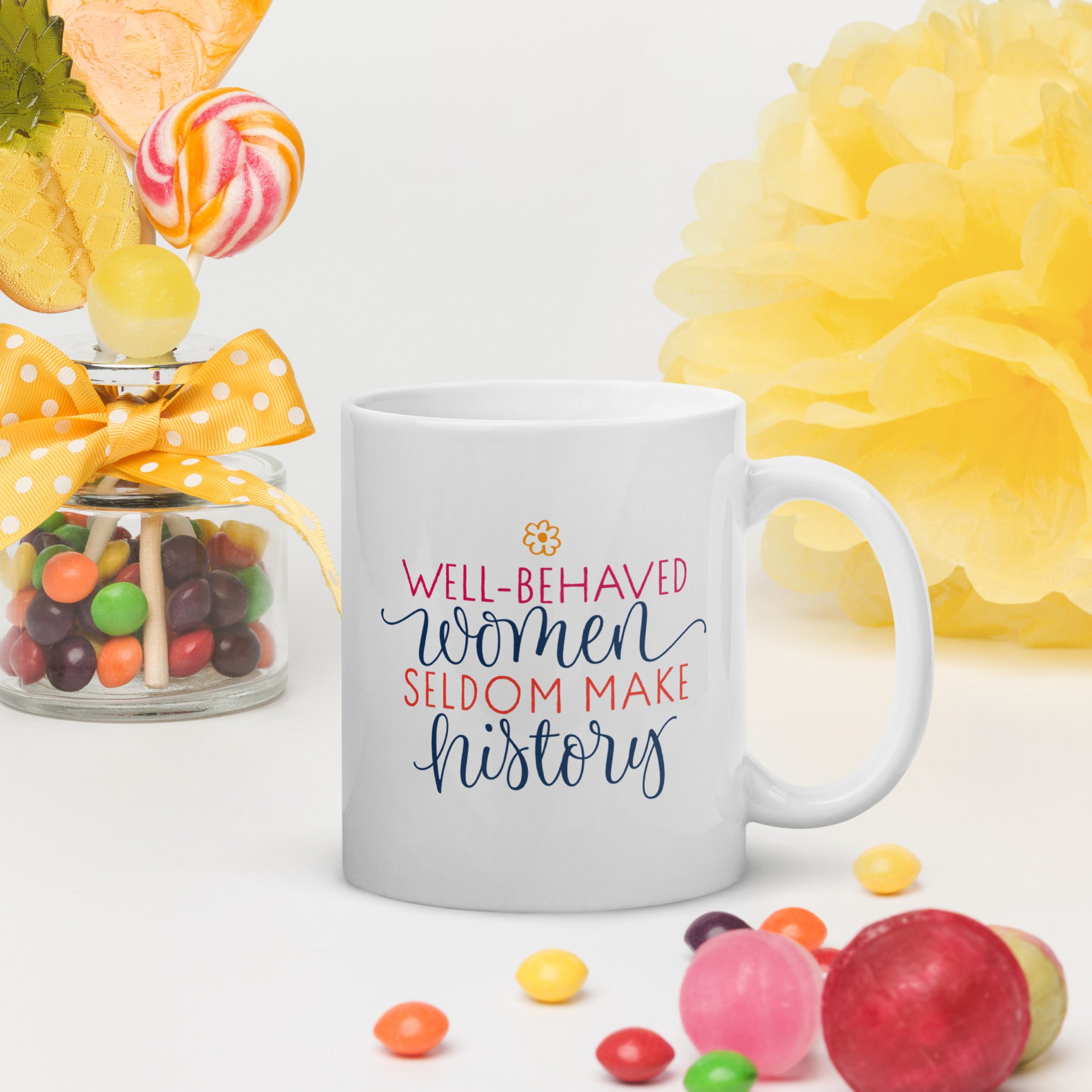 White mug with motivational quote surrounded by colorful candies and flowers on a light background