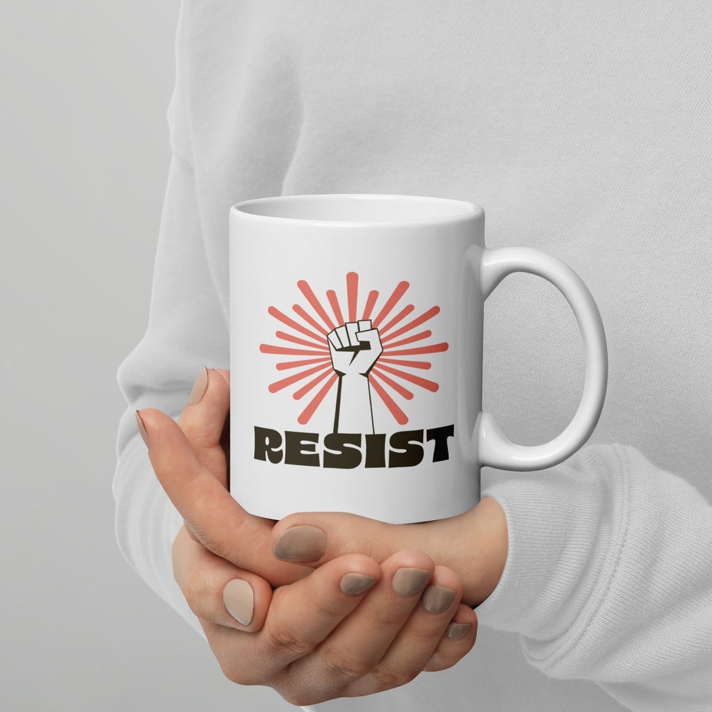 Person holding coffee mug with handle on right. Mugh has a raised fist graphic in front of a coral burst, and bold retro-styled text that says "resist."
