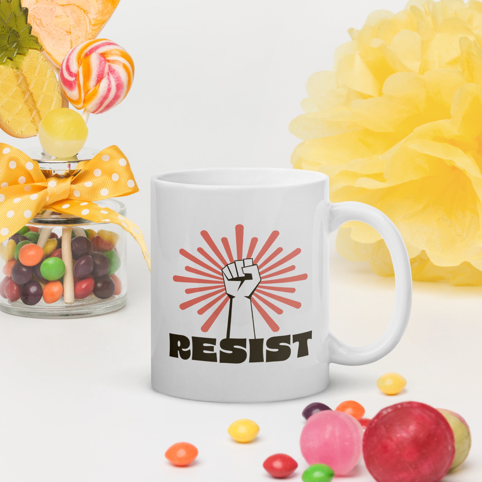 Coffee mug in a scene with brightly colored candy. Mug has a raised fist graphic in front of a coral burst, and bold retro-styled text that says "resist."