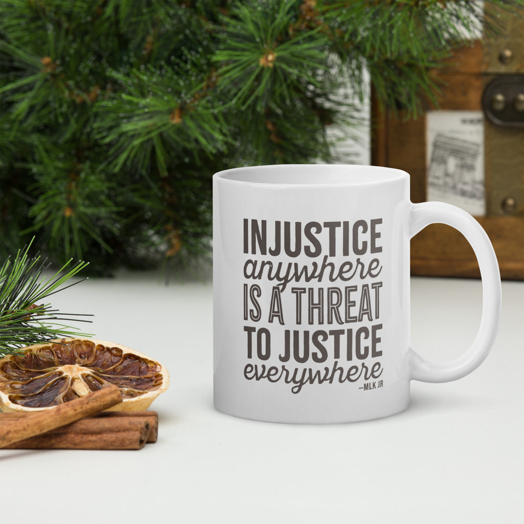 White ceramic mug with bold text graphic reading “injustice anywhere is a threat to justice everywhere,” a quote from MLK Junior from Letters from Birmingham Jail, on a tabletop with forest decorations.