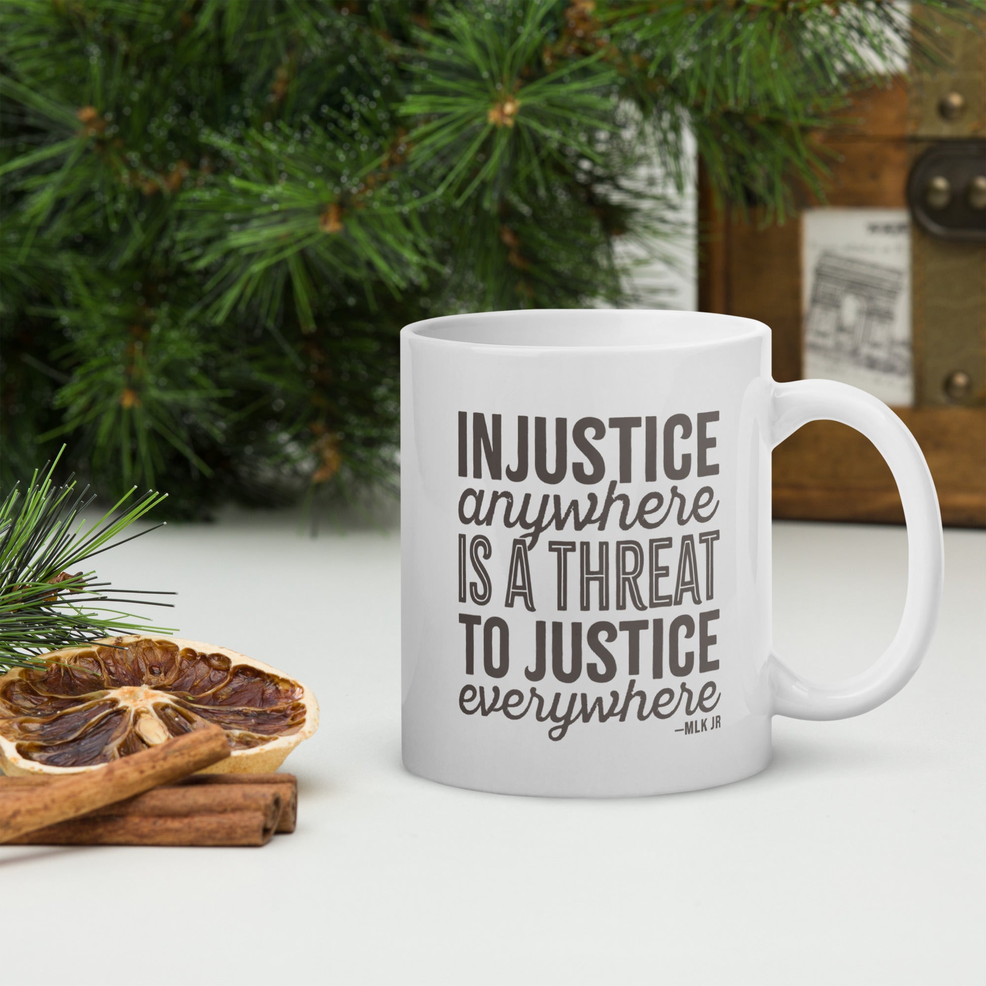 White ceramic mug with bold text graphic reading “injustice anywhere is a threat to justice everywhere,” a quote from MLK Junior from Letters from Birmingham Jail, on a tabletop with forest decorations.