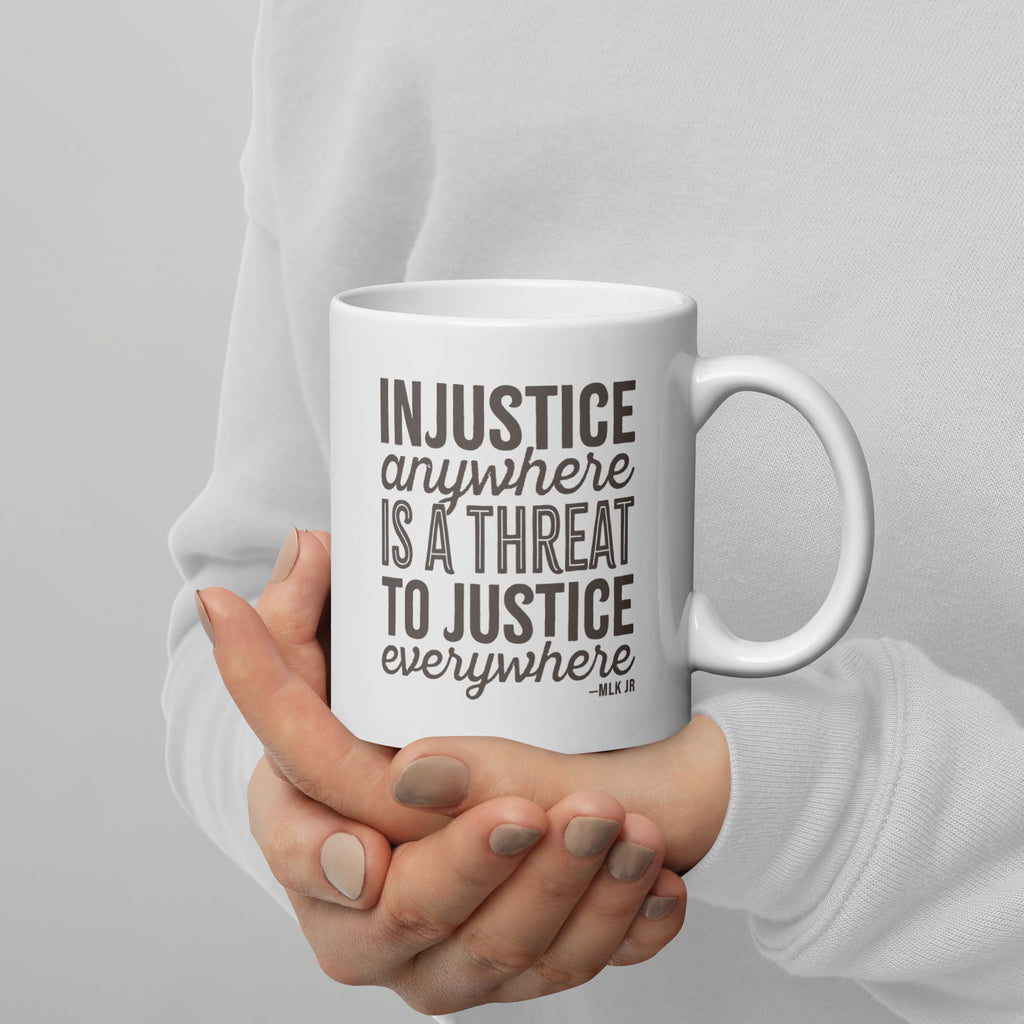 Person holding white ceramic mug showing right side with bold text graphic reading “injustice anywhere is a threat to justice everywhere,” a quote from MLK Junior from Letters from Birmingham Jail.