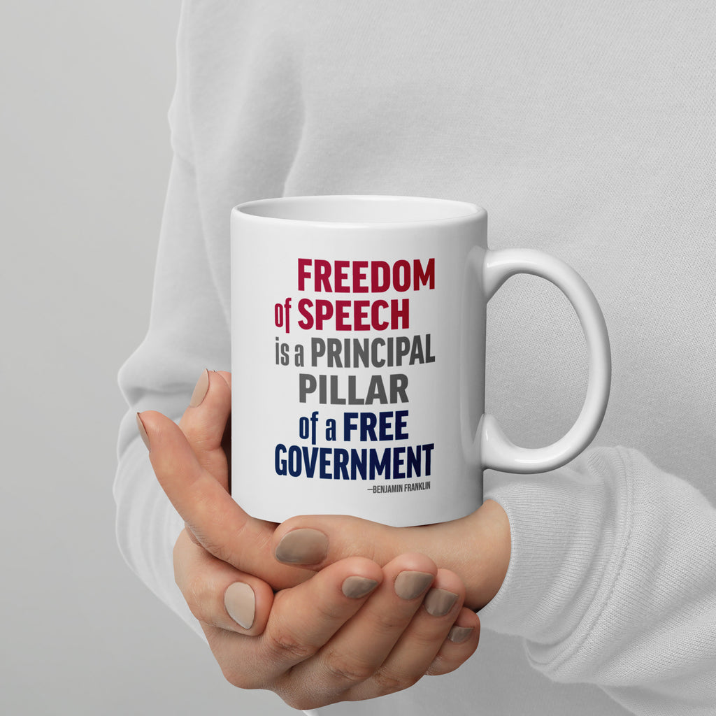 Person holding white ceramic mug with bold patriotic red, blue, and grey text that reads "Freedom of speech is a principal pillar of a free government. – Ben Franklin."