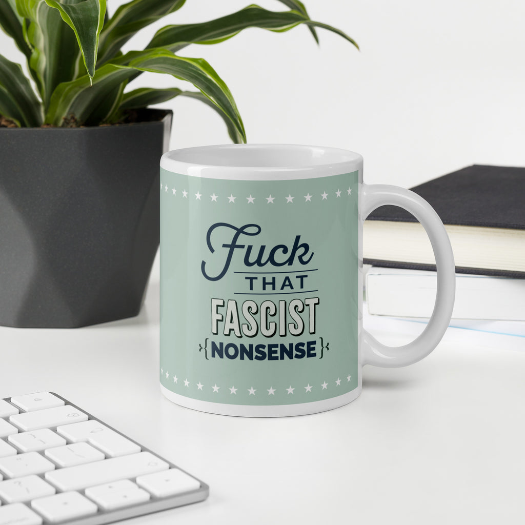 Ceramic mug with graphic on mint green background and navy styled text block that reads "fuck that fascist nonsense." Mug is on a desk with a plant and a keyboard.