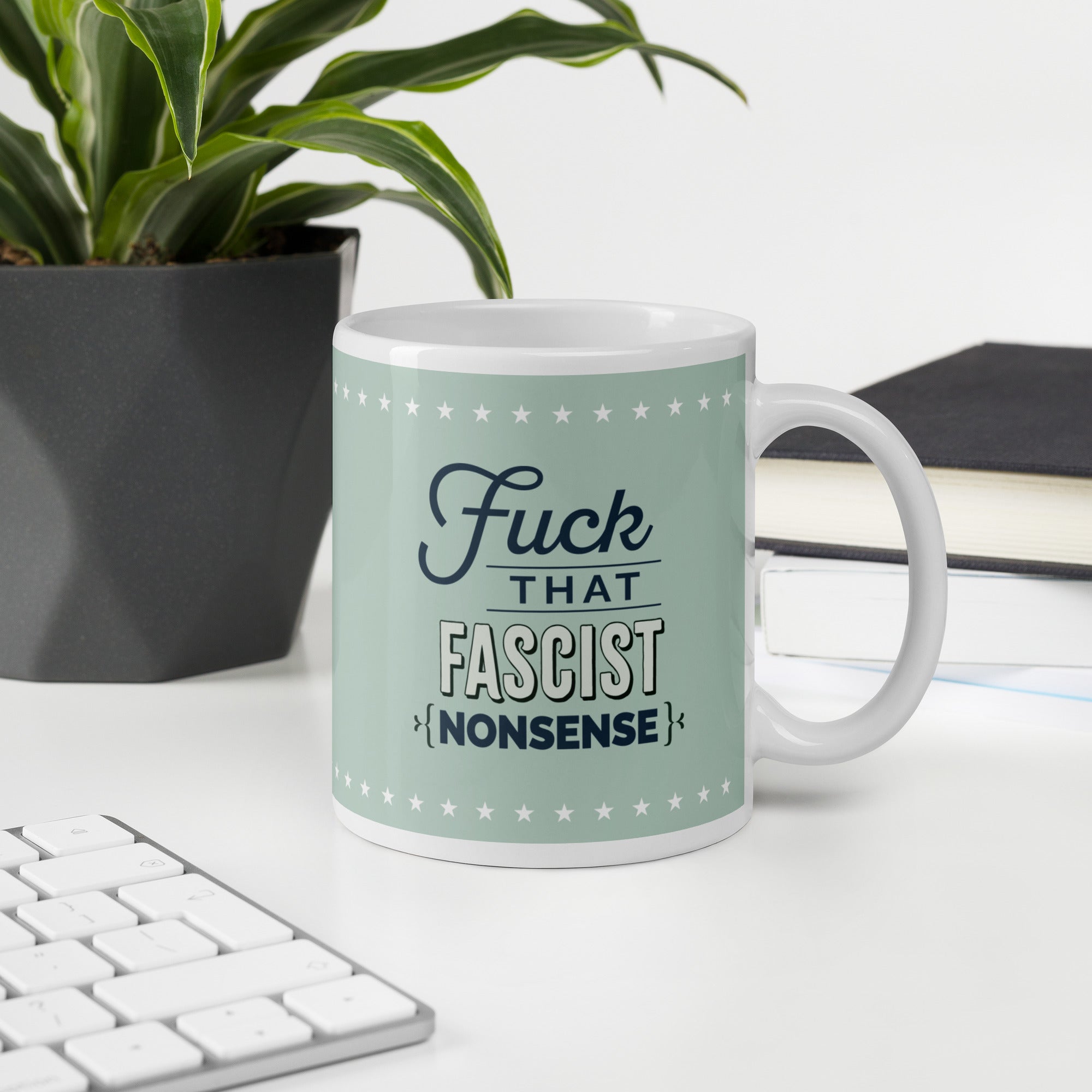 Ceramic mug with graphic on mint green background and navy styled text block that reads "fuck that fascist nonsense." Mug is on a desk with a plant and a keyboard.