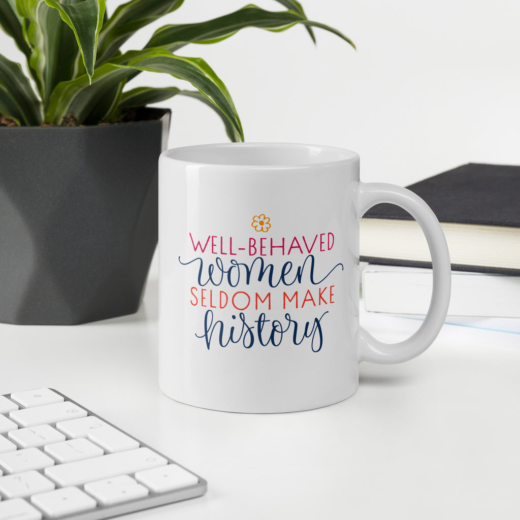 White mug with motivational quote that reads "well-behaved women seldom make history" on a desk with keyboard, plant, and notebook.