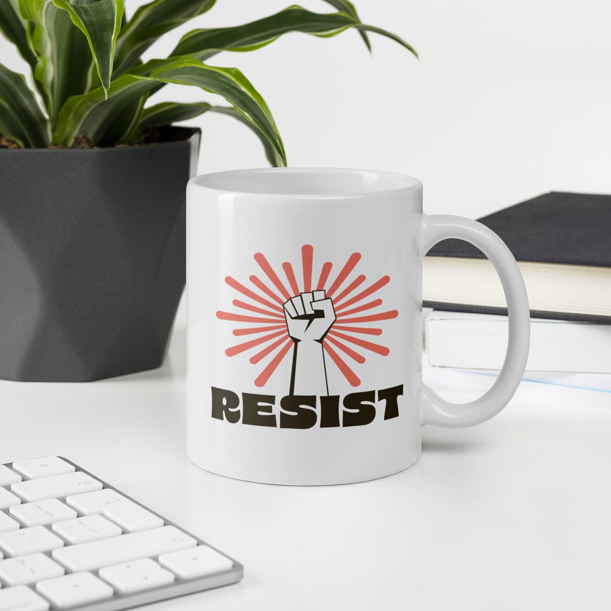 Coffee mug in an office desk scene. Mug has a raised fist graphic in front of a coral burst, and bold retro-styled text that says "resist."