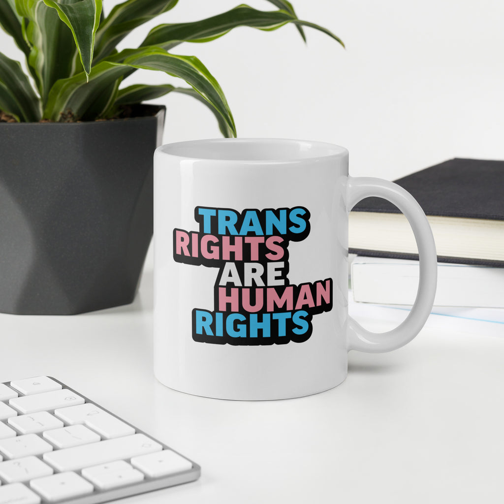 White ceramic mug with bold text “Trans Rights Are Human Rights” in blue, pink, and black colors representing the transgender pride flag, on a desk with a keyboard and a plant.