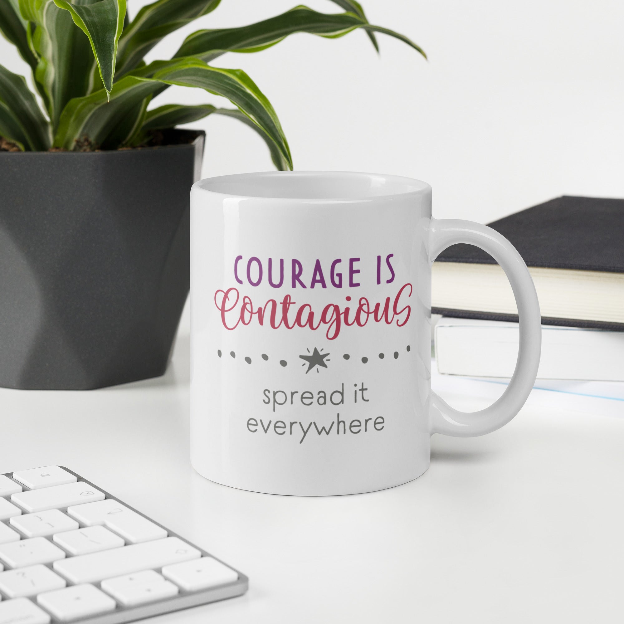 Mug with styled text graphic that reads "courage is contagious – spread it everywhere", on a desk with plant and keyboard.