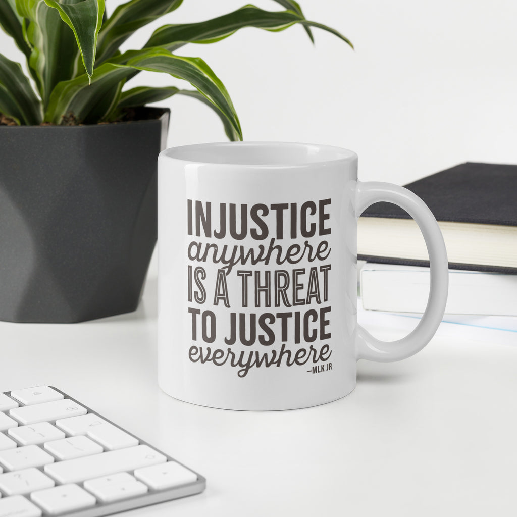 11 ounce white ceramic mug with bold text graphic reading “injustice anywhere is a threat to justice everywhere,” a quote from MLK Junior from Letters from Birmingham Jail, on a desk with a plant and a keyboard.