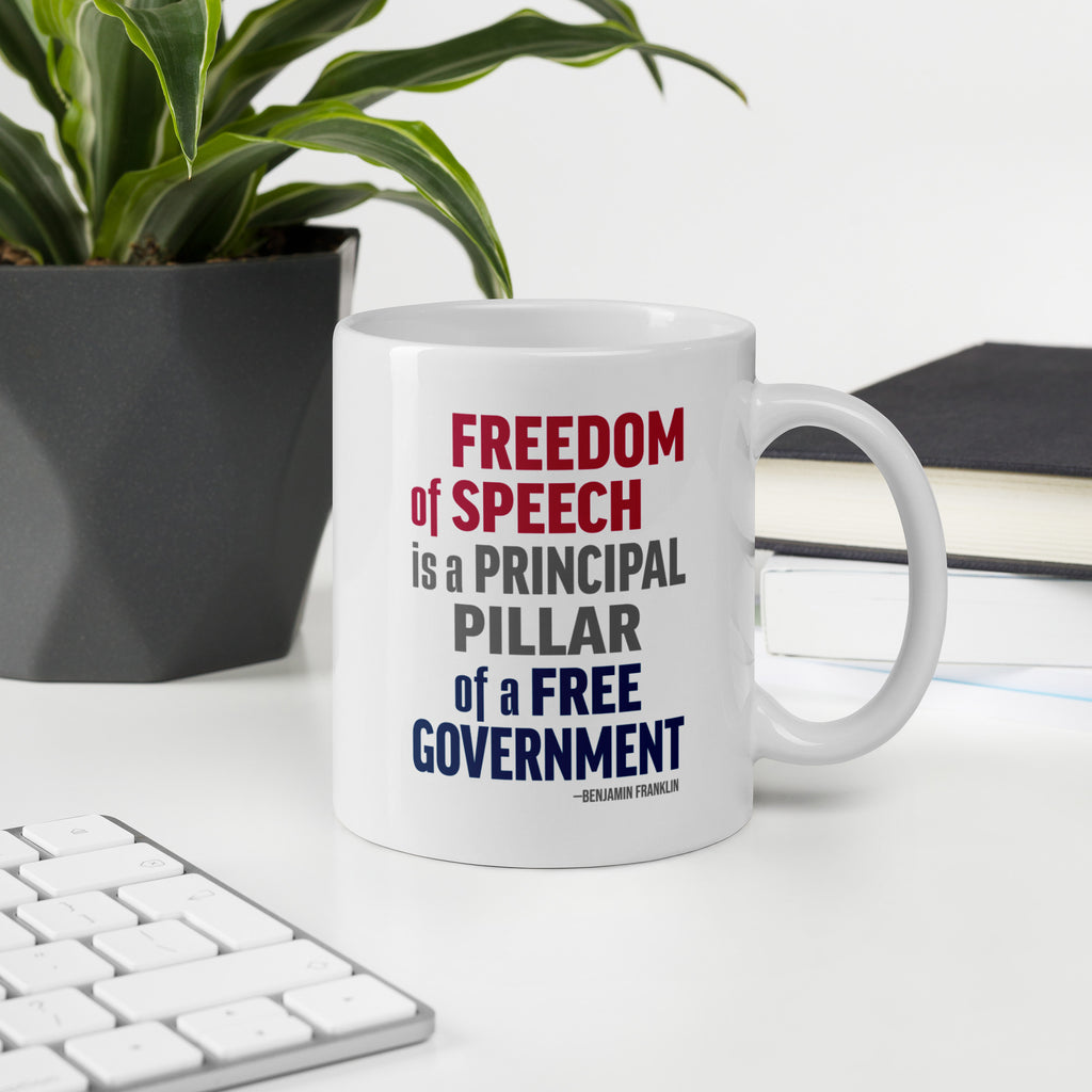 White ceramic mug with bold patriotic red, blue, and grey text that reads "Freedom of speech is a principal pillar of a free government. – Ben Franklin." On a desk with a plant, book, and a keyboard.