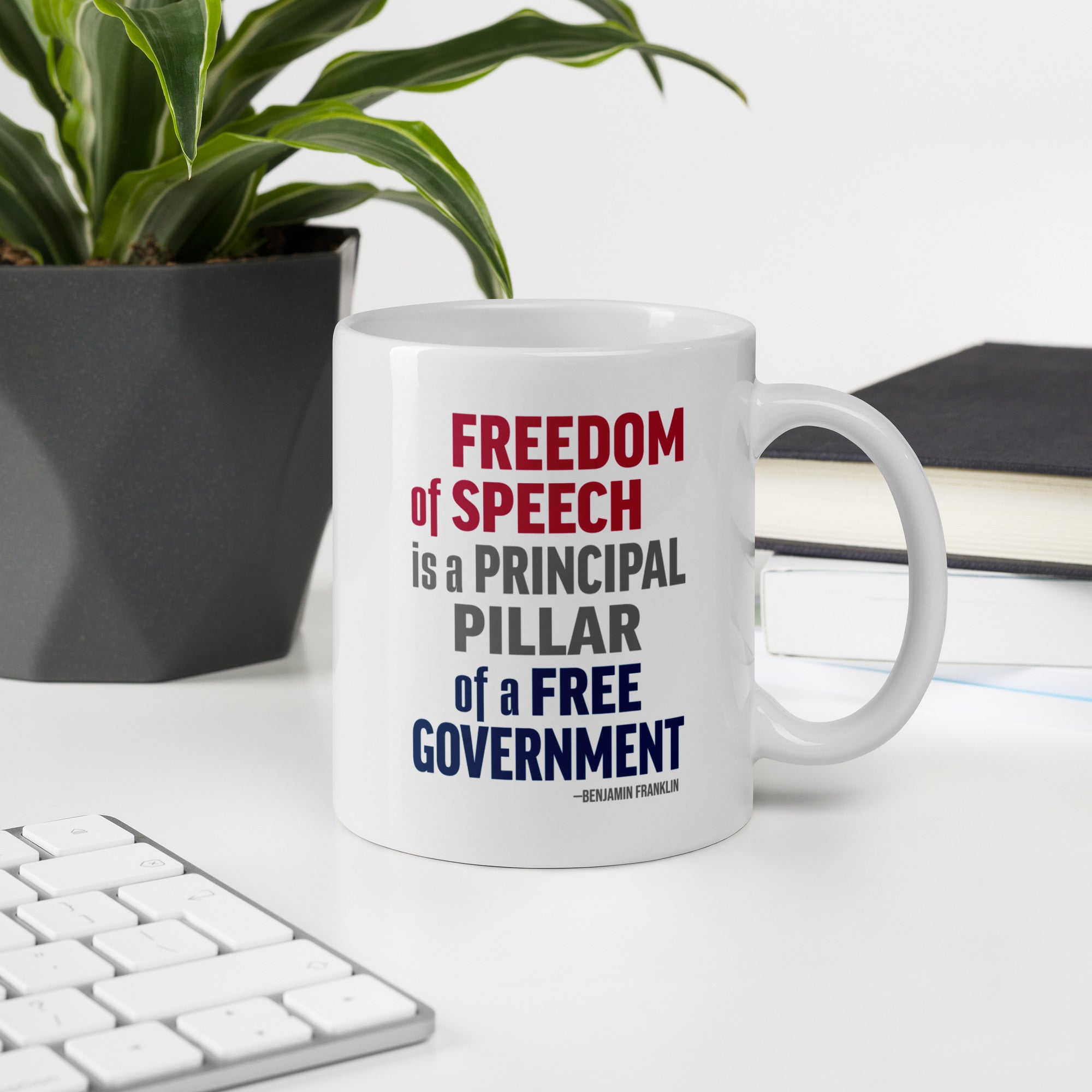 White ceramic mug with bold patriotic red, blue, and grey text that reads "Freedom of speech is a principal pillar of a free government. – Ben Franklin." On a desk with a plant, book, and a keyboard.