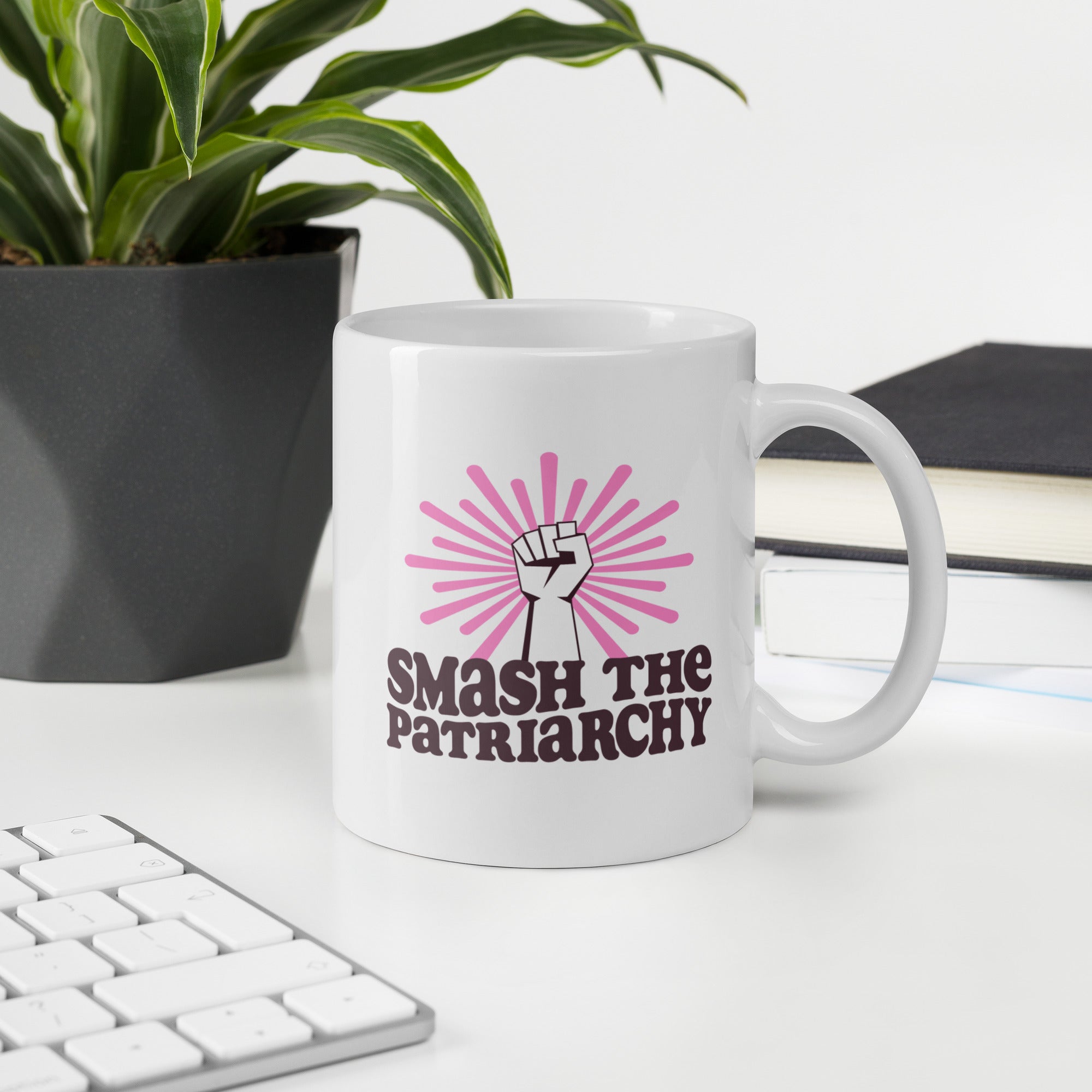 Coffee mug that says "smash the patriarchy" below a raised fist centered in front of pink rays, on a desk with a keyboard, plant, and book.