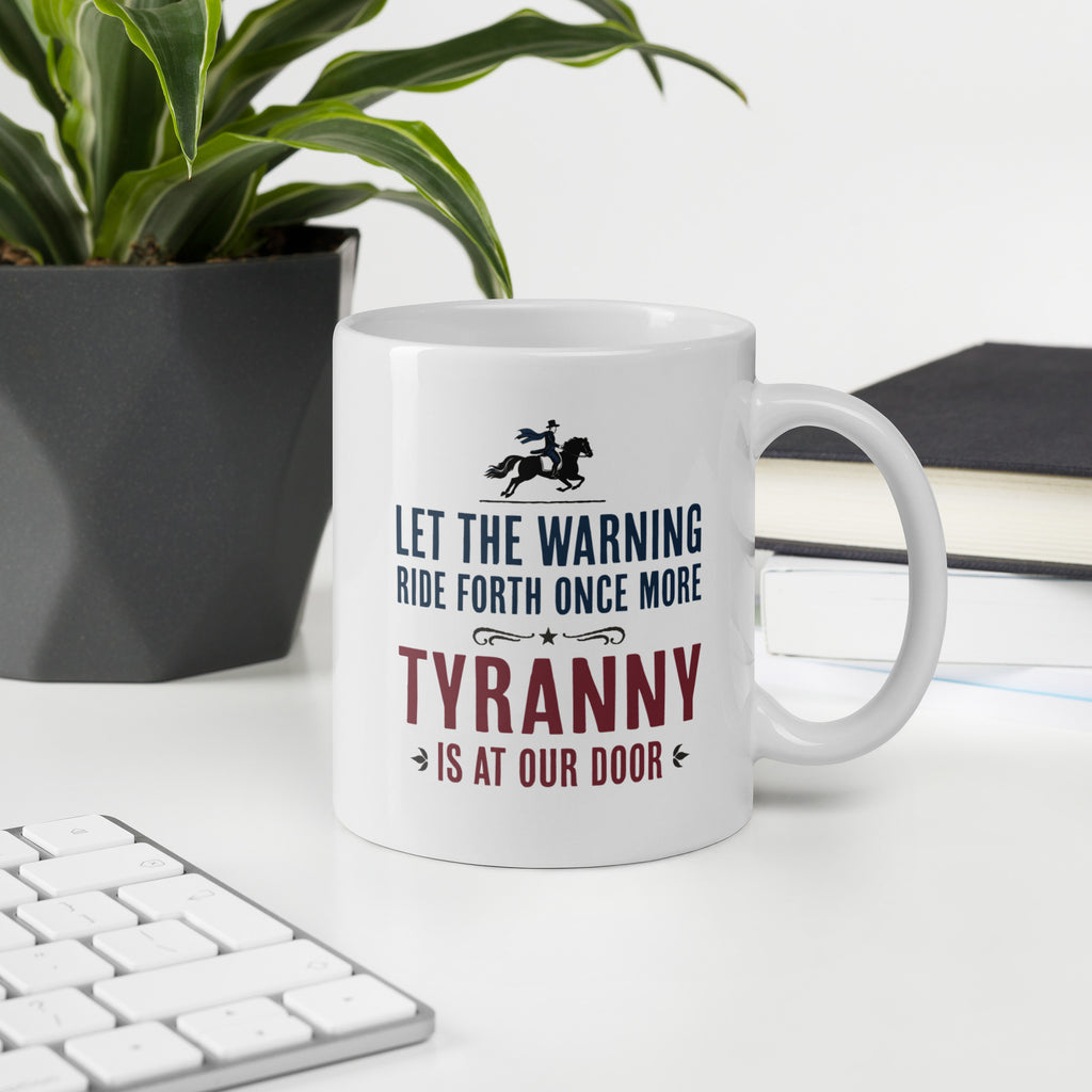 White ceramic coffee mug with a historical-style design reading “Let the warning ride forth once more — Tyranny is at our door,” on a desk with a keyboard, book, and plant.