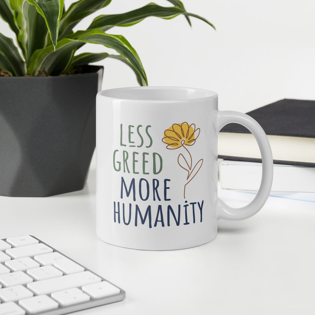 White ceramic mug featuring the phrase “Less Greed More Humanity” in hand-lettered typography with green and blue text alongside a simple yellow flower illustration, on a desk with a keyboard, book, and plant.