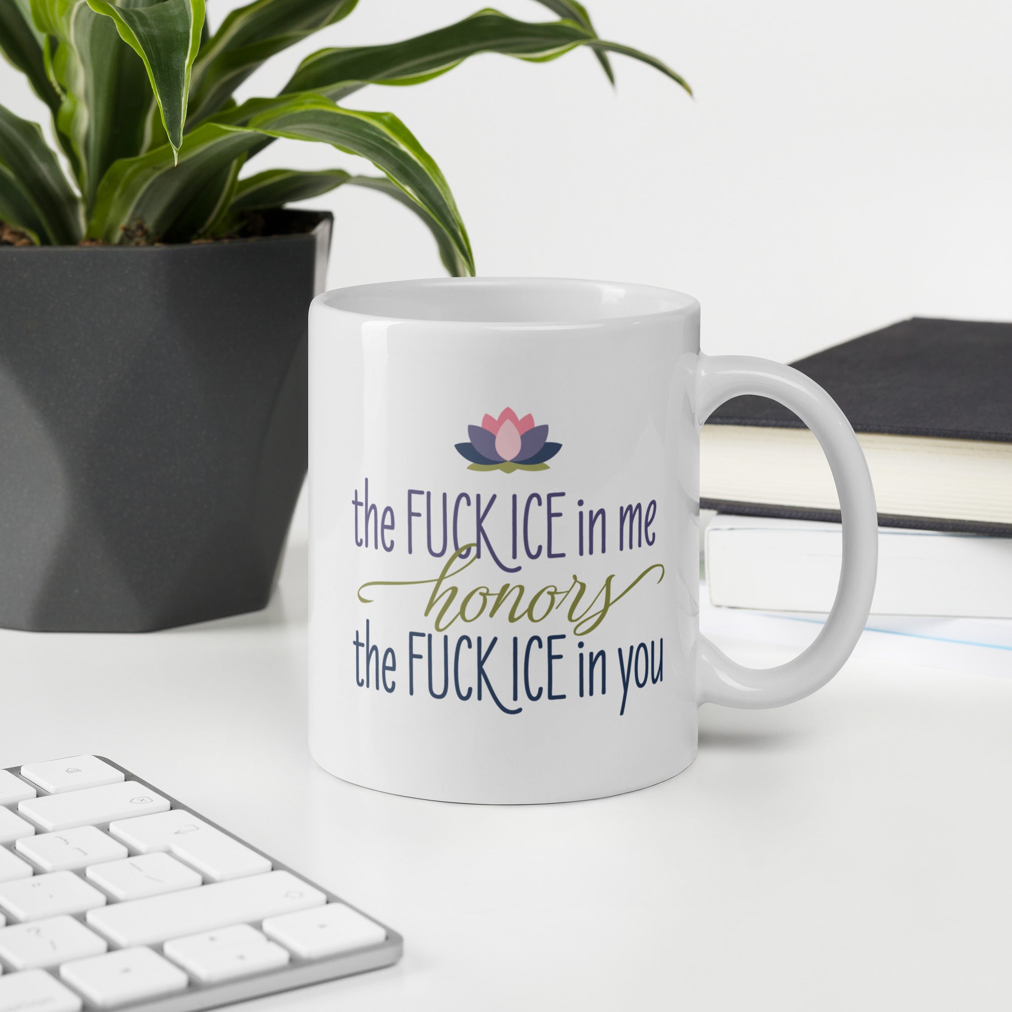 White ceramic coffee mug featuring the phrase "The Fck ICE in Me Honors the Fck ICE in You" in decorative typography with a lotus flower above, on a desk with a keyboard, book, and plant.