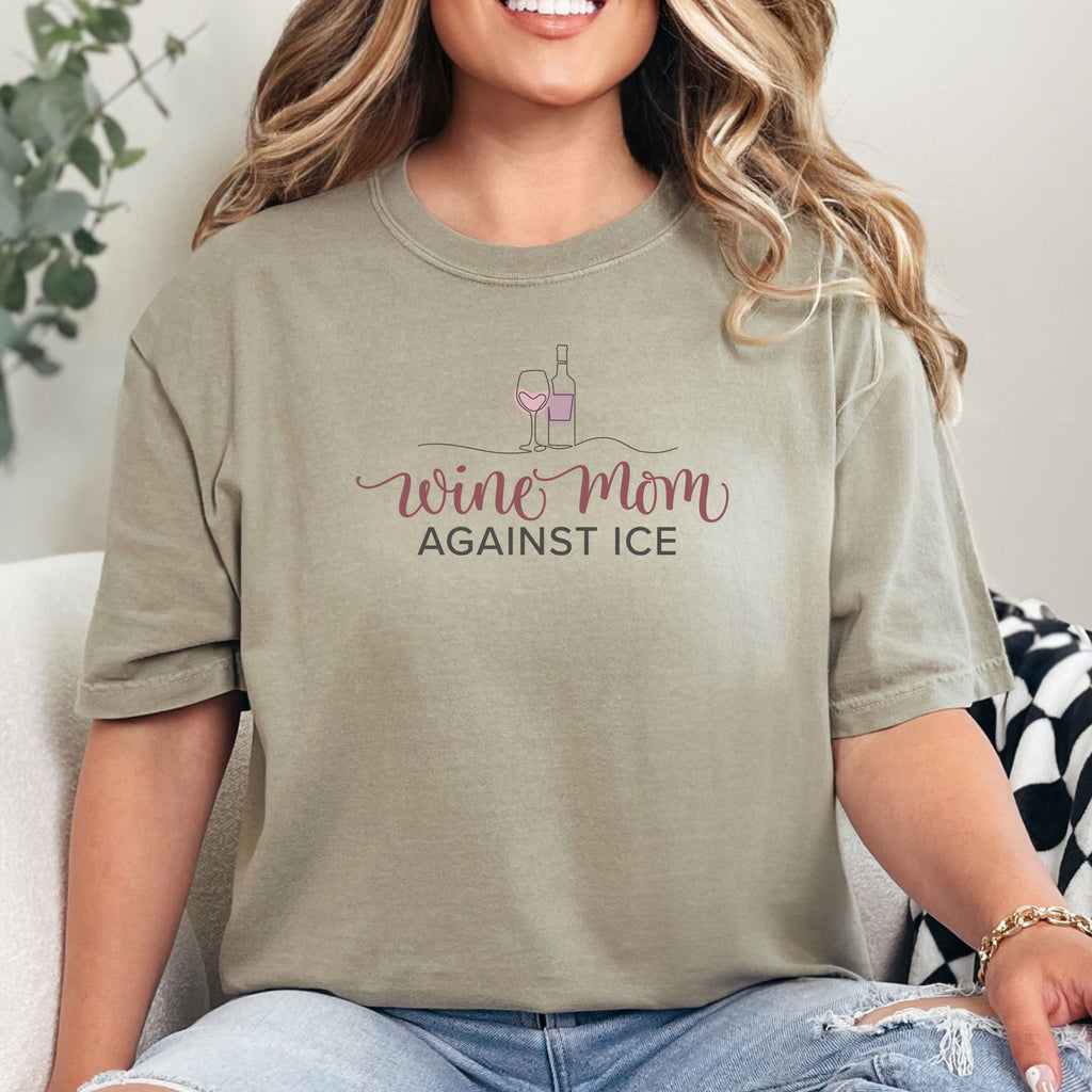 Female model wearing khaki t-shirt featuring the text “Wine Mom Against ICE” with a minimalist illustration of a wine bottle and glass above the lettering.