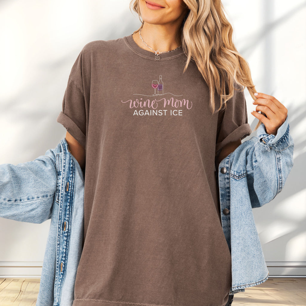 Female model wearing espresso t-shirt featuring the text “Wine Mom Against ICE” with a minimalist illustration of a wine bottle and glass above the lettering.