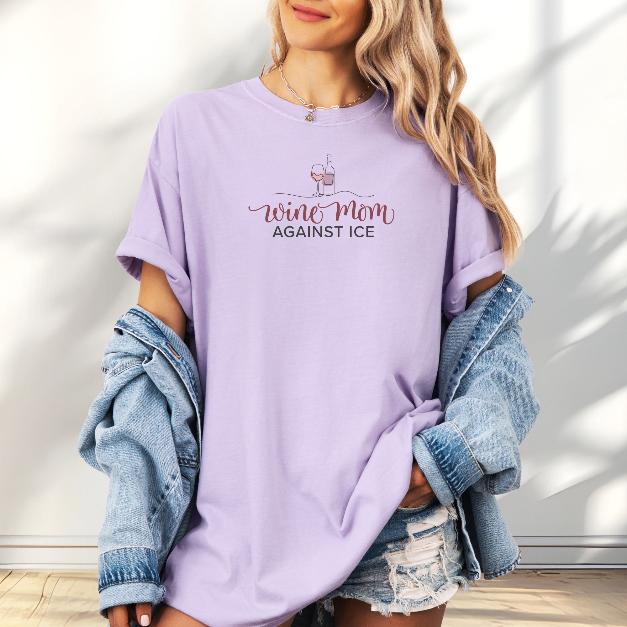 Female model wearing oversized orchid t-shirt featuring the text “Wine Mom Against ICE” with a minimalist illustration of a wine bottle and glass above the lettering.