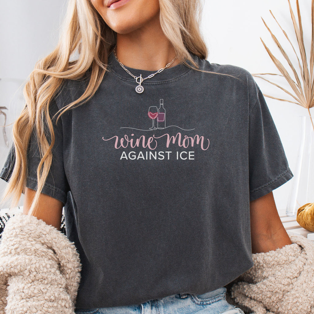 Female model wearing pepper t-shirt featuring the text “Wine Mom Against ICE” with a minimalist illustration of a wine bottle and glass above the lettering.