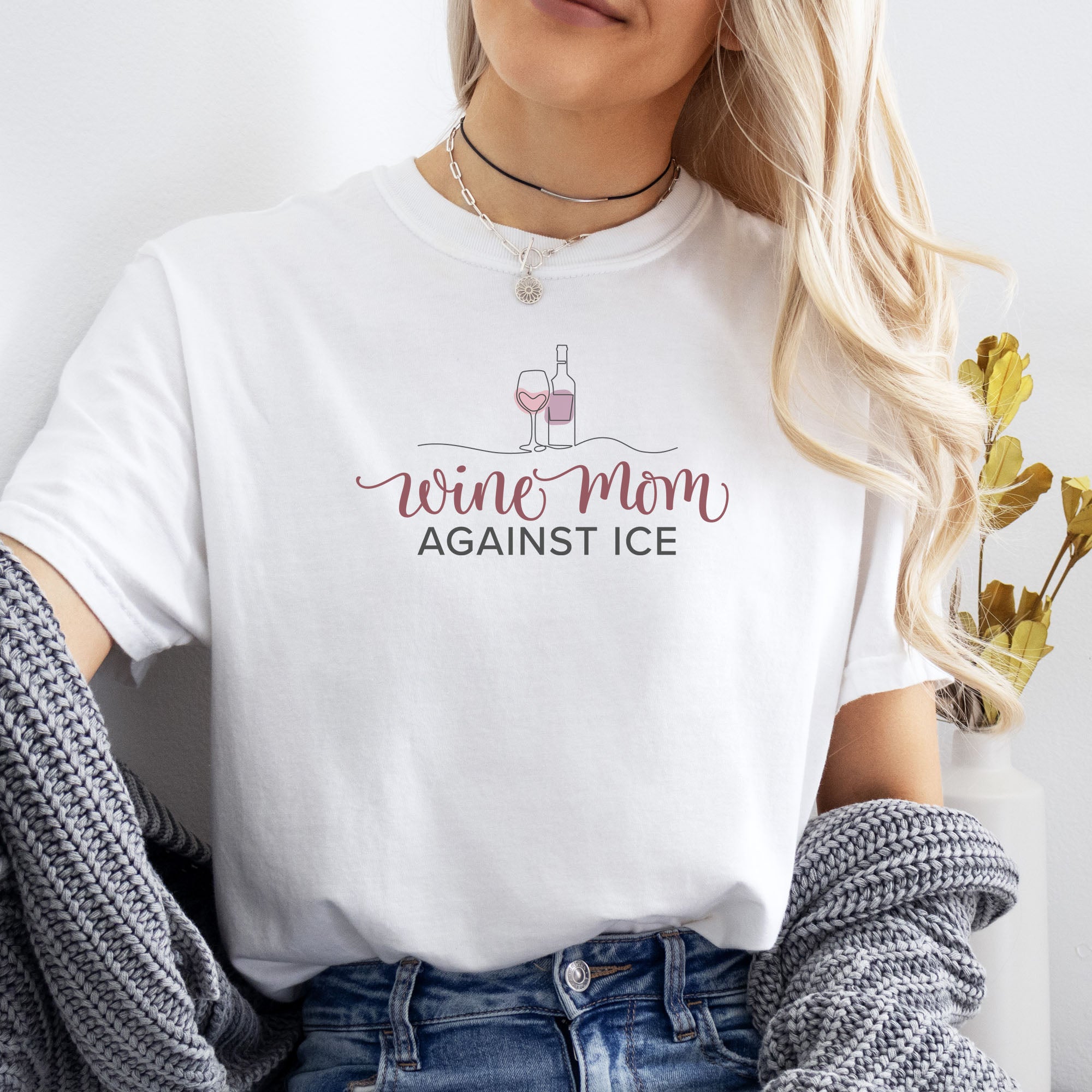 Female model wearing white t-shirt featuring the text “Wine Mom Against ICE” with a minimalist illustration of a wine bottle and glass above the lettering.